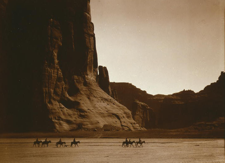 Photograph of a number of people on horseback roaming the an old west landscape.