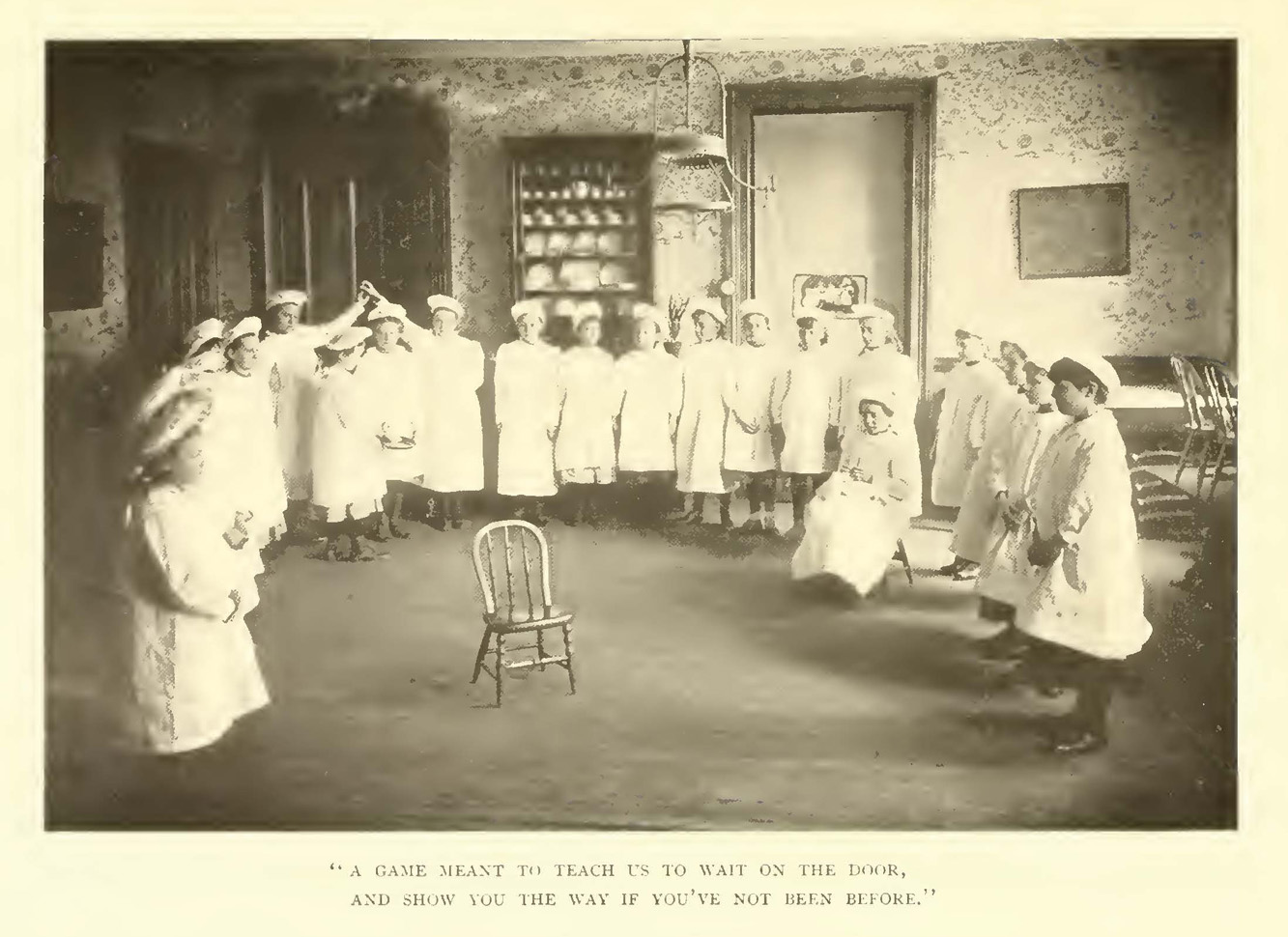 A group of girls in white smocks and caps stand around a chair in a 1901 classroom.