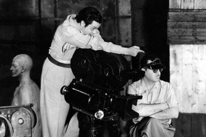 Black and white photo of a director behind a large film camera and pointing, while another man sits in a chair next to it with his arms crossed wearing sunglasses and a visor.