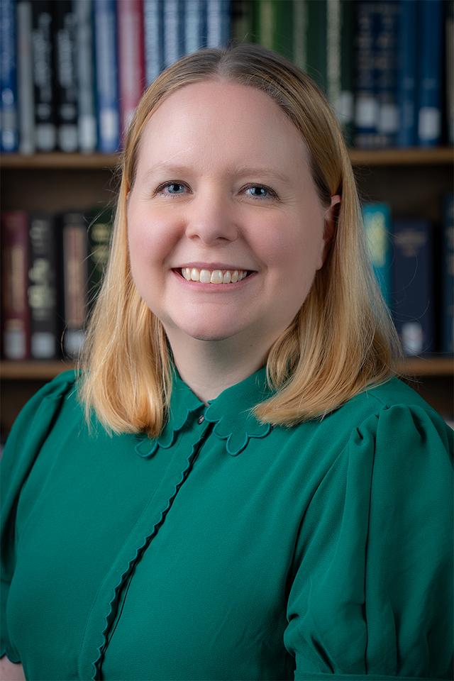Photograph of Joanna Thielen, a white woman with short blond hair wearing a green shirt, seated in front of a bookcase.
