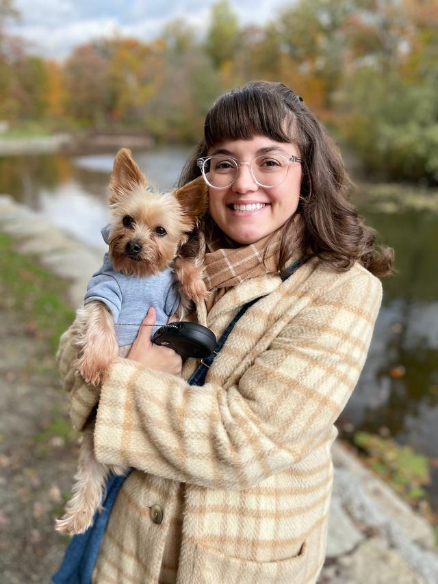 White woman with shoulder length brown hair, wearing clear frame glasses, a tan and beige plaid coat, and holding a small terrier dog in grey sweatshirt. 