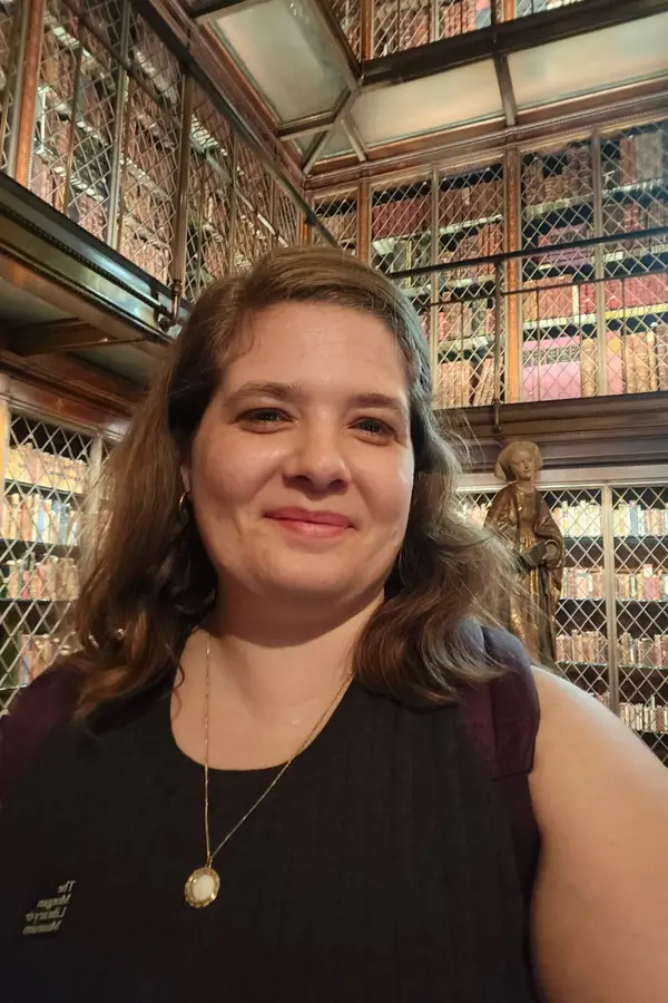 A white woman with shoulder length blonde hair posing in front of a massive wall of rare books in the Morgan library in NYC