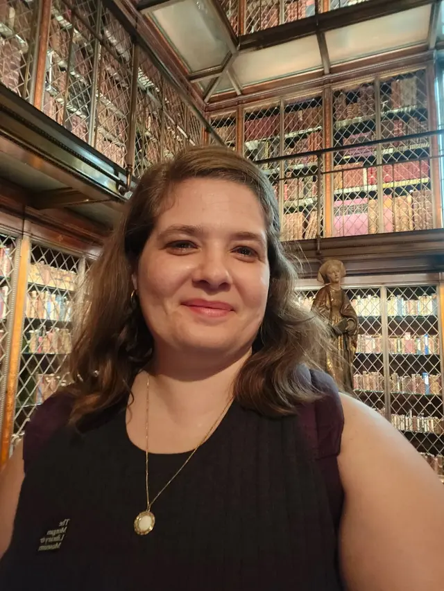 A white woman with shoulder length blonde hair posing in front of a massive wall of rare books in the Morgan library in NYC