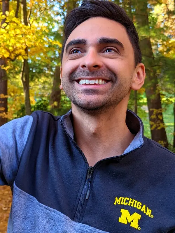 Josh wearing a University of Michigan sweater and smiling while standing outdoors with fall-colored trees in the background