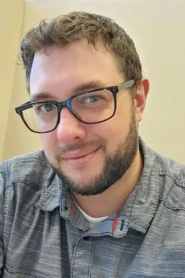 Photograph of Erik LaCoss, a white male with short brown hair, brown glasses in a grey button down shirt.