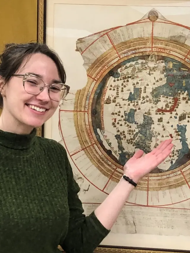A white woman in a green turtleneck gestures behind her towards a circular map of the world from 1542.