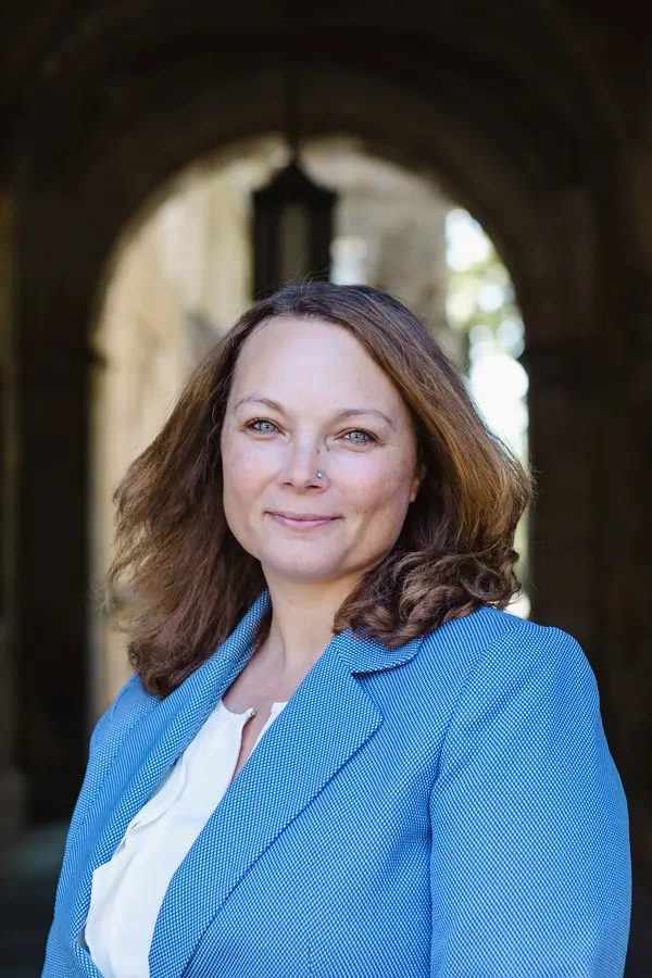 The picture is located near an archway to the Law Quadrangle at the University of Michigan's Ann Arbor campus. I am a white female with shoulder-length light brown hair, and I have blue eyes. I am wearing a light blue suit jacket with a white shirt underneath.