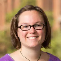 Photograph of Sarah Barbrow, a white person with short brown hair wearing clear glasses, with blurred trees in the background