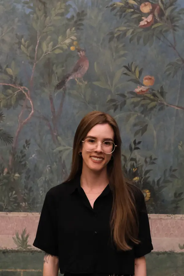 Photo of Rachel, a white woman with long brown hair and glasses, standing in front of a Roman fresco and smiling.