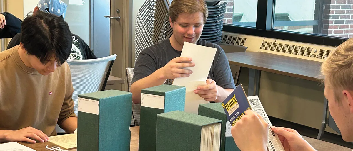 Three students reading journals at a table with four green journal boxes.