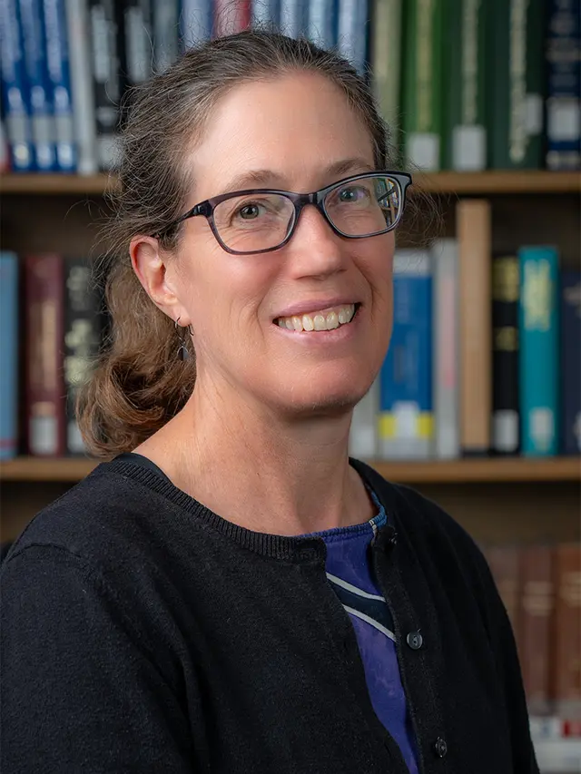 A photo of a white woman with a ponytail and glasses, wearing a multicolored dress and a black sweater.