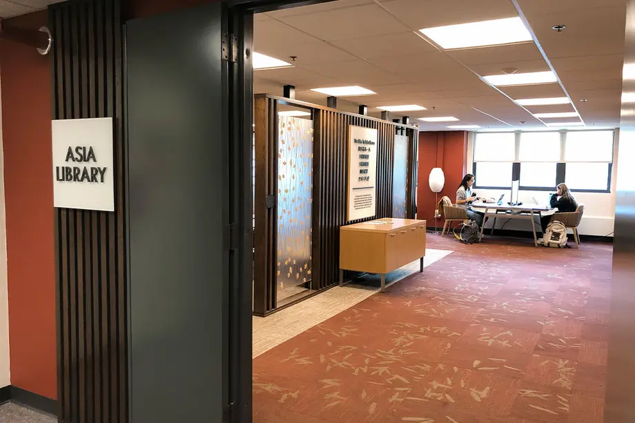 Open door into the Asia Library space with bright light shining through the windows. Asia Library sign to the left of the door with vertical wooden slats behind it.