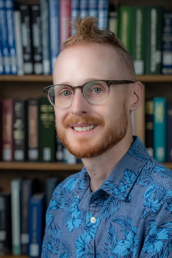 A photo of a smiling person with acetate-framed glasses, a short beard, and red hair styled in a mohawk. He is wearing a blue floral-patterned shirt.