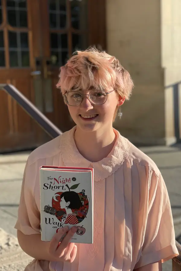 A white person with short pink hair, large round glasses, and a pink button up shirt smiles and holds up the book The Night is Short, Walk on Girl