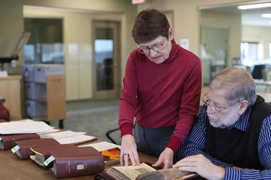 Woman standing next to a man at a desk, with both looking intently at the book open on the desk.