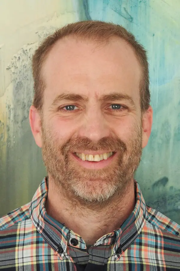A smiling, white, older male with short blond hair and beard, wearing a chequered shirt, standing against a multicolor abstract background.