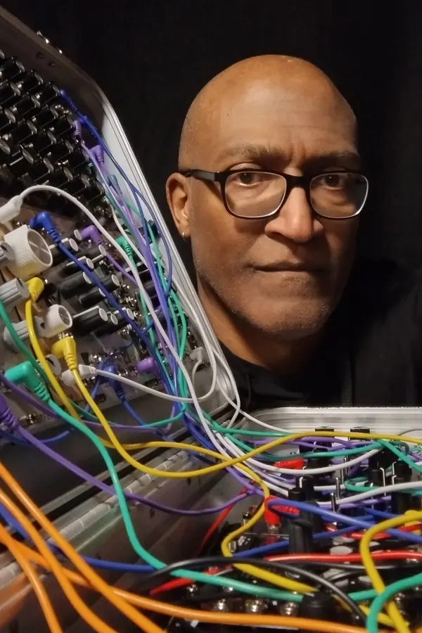 A person with glasses poses next to a modular synthesizer filled with colorful cables, set against a dark background.