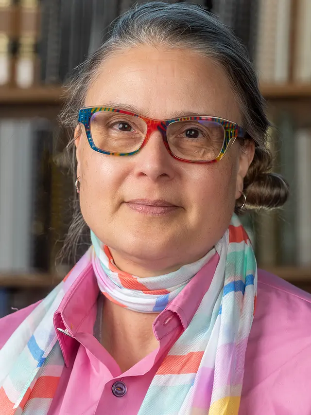 Photo of Marilène "Mars" De Ritis - a woman with greying hair and multi-colored eyeglass frames wearing a magenta shirt and light-weight scarf in front of Reference Reading Room bookshelf