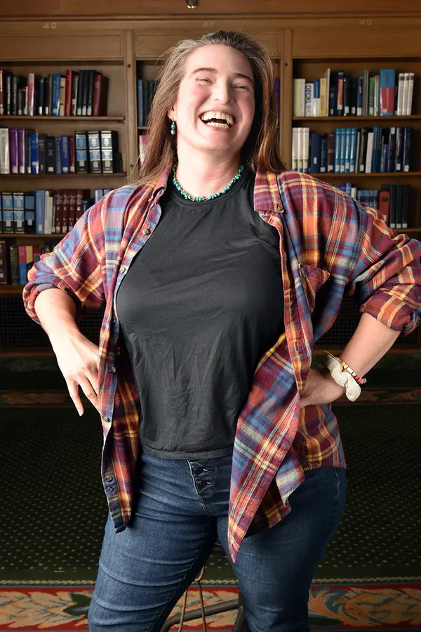 Laughing woman in colorful plaid with hands on hips stands before full bookcases.