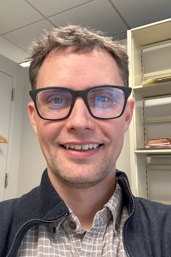 A smiling, bespectacled middle-aged white man with short brown hair wearing a checkered shirt and blue sweater in front of a bookcase.