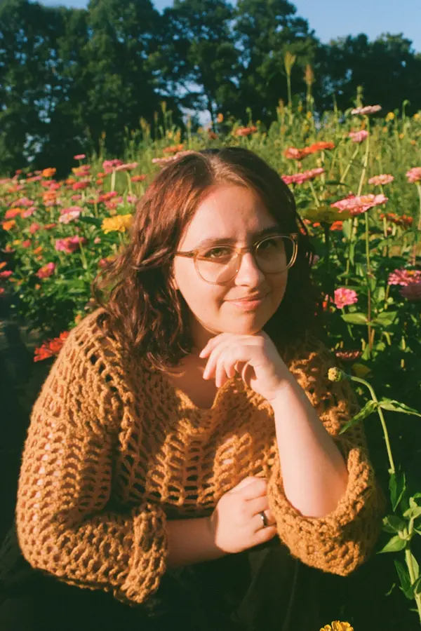Young adult woman kneeling in a field of flowers during daytime and making eye contact with the viewer. She is wearing a yellow crocheted shirt and resting her head on her hand.