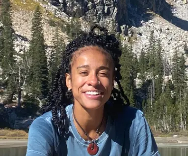 An AFAB person with long black braids is smiling in front of an alpine lake and mountains