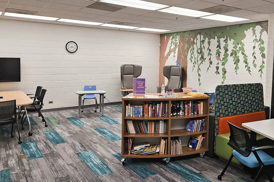 A wall with a jungle mural, high back chairs, child-size desk, little free library lending book shelves, and table with a shared display.