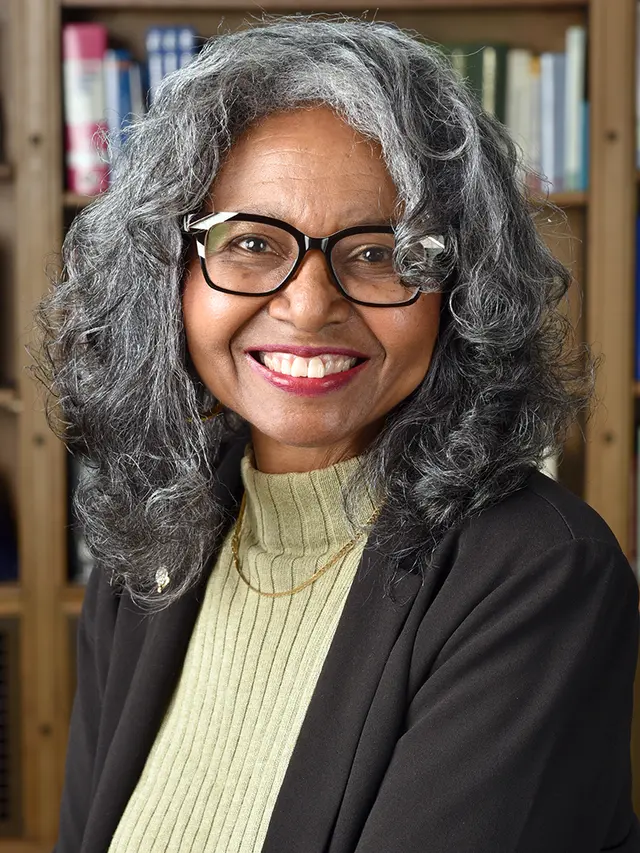 Black woman with salt & pepper curls, black and white glasses, black blazer pale olive sweater.
