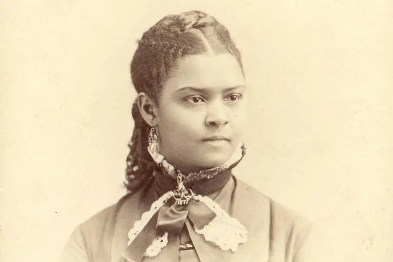 Monochrome seated portrait photograph of an African American woman with braids in her hair and high collared dress.