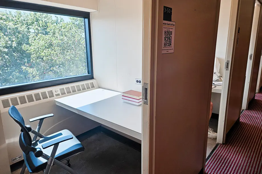 Chair and work surface in a study carrel with a window and trees outside, there's a sign outside the entrance that it's bookable.