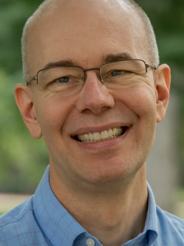 Photograph of Scott Dennis, a white man with a mostly bald head and very short gray hair on the sides, wearing clear glasses, a blue shirt, and a big smile in front of green foliage.