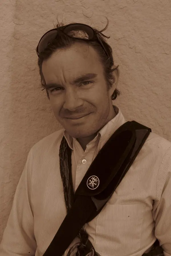 Photograph of Ben Howell, white male with short hair and a light button down shirt, standing outside against a light rock wall.