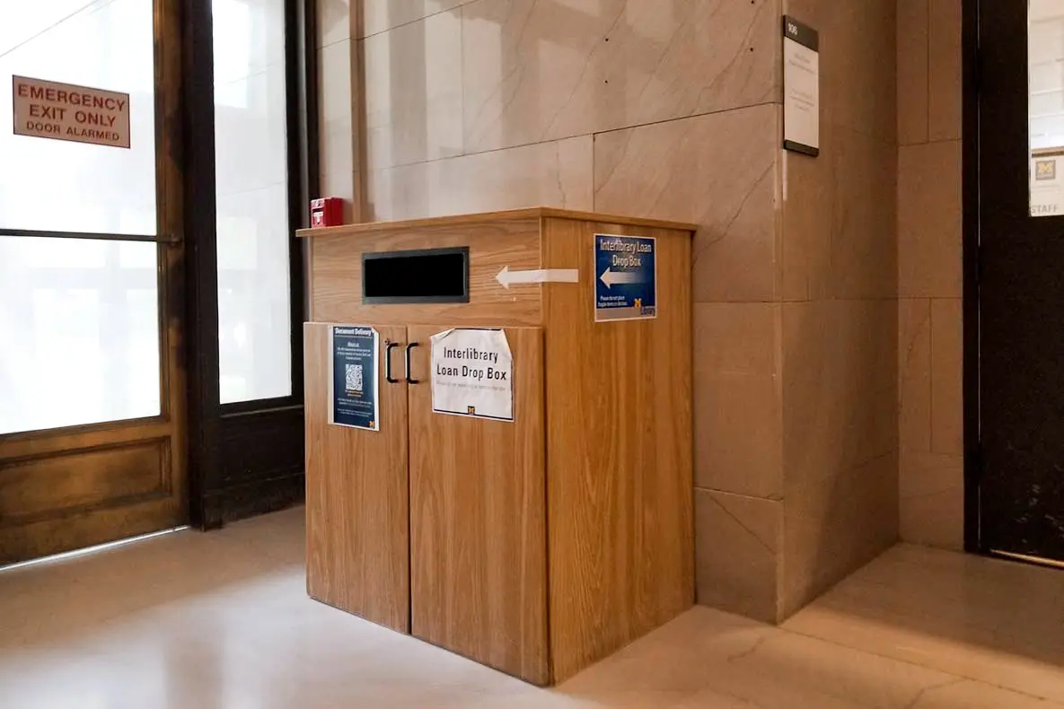 Wood interlibrary loan dropbox with slots and signage, emergency exit to the left.