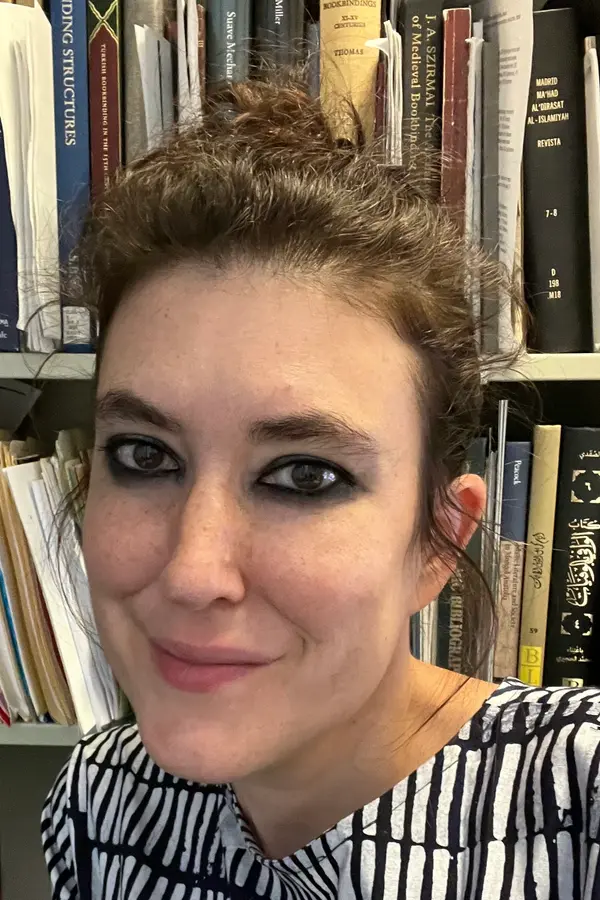 headshot of Evyn Kropf, a person with light skin, wavy dark hair, dark eyebrows and dark eyes, in front of a bookcase overflowing with books