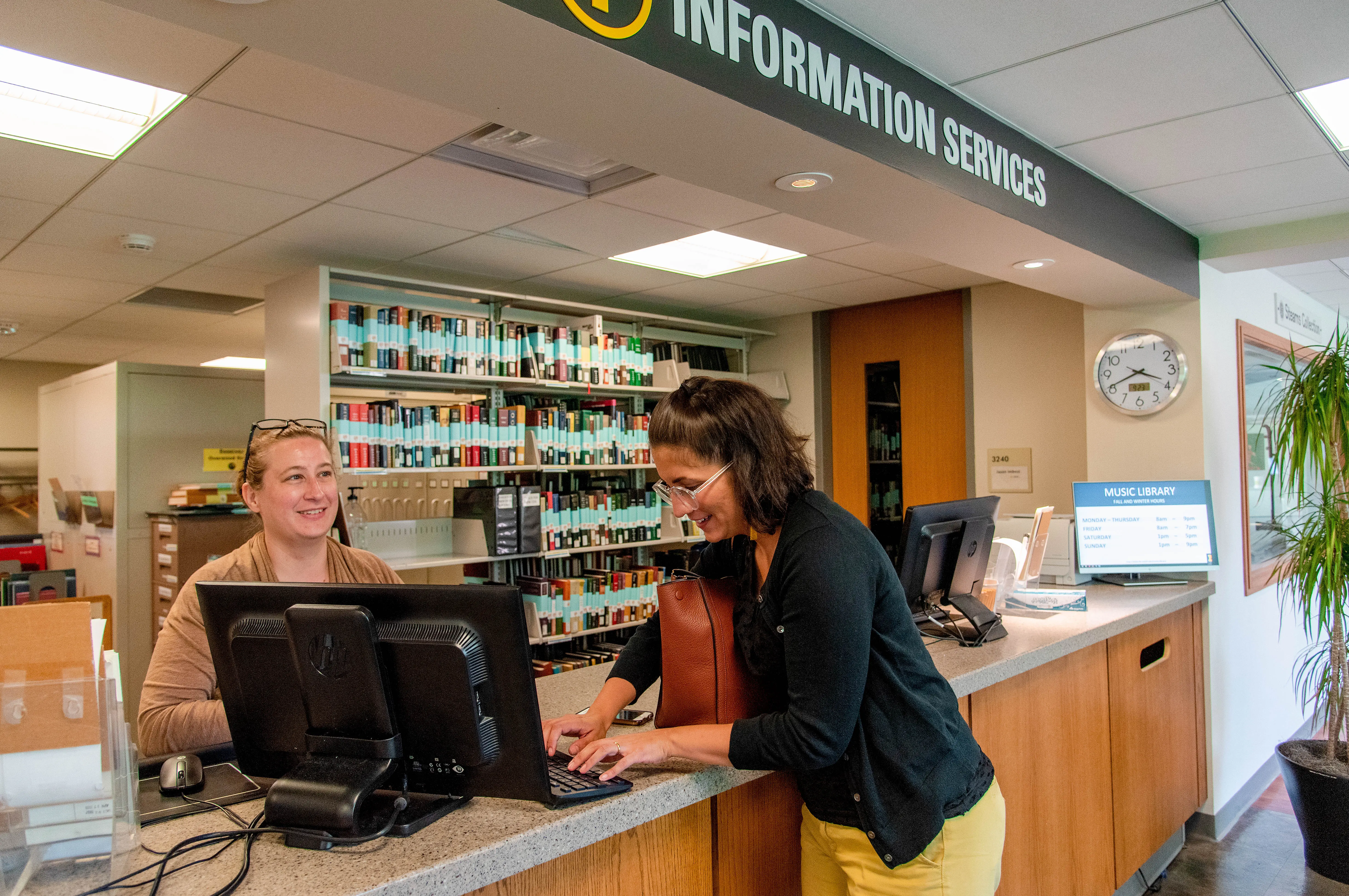 Person behind a computer at the Music Library Information Service Desk, talking to a student.