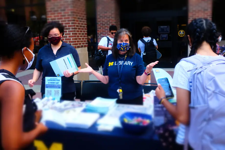 A university librarian in a blue T-shirt talks with students at a table outside the Shapiro Library during the fall semester's FestiFall event.