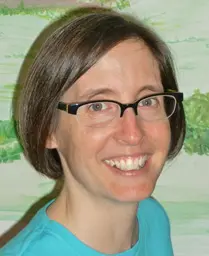 Photograph of Beth Snyder, a white woman with glasses and short brown hair wearing clear glasses and a turquoise top, in front of painted mural.