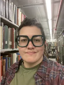 A picture of Shannon Moreno. A person with short dark hair, large square green glasses, wearing an olive green shirt with burgundy flannel, and standing in front of books.