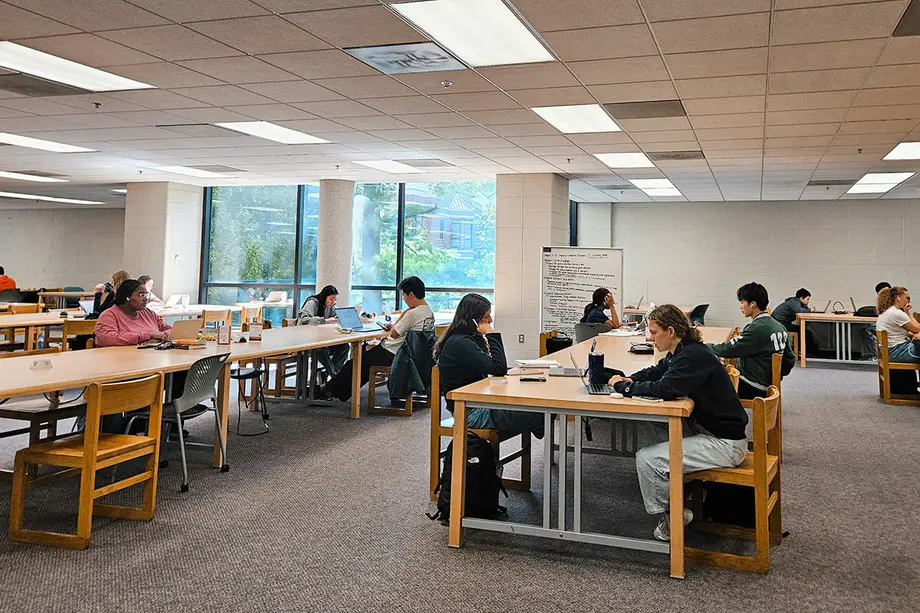 Students studying at long rectangular tables with a movable whiteboard in view and a window in the background.
