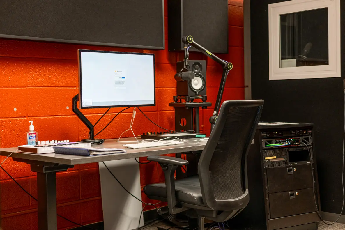 A desktop computer with attached equipment and soundproof booth window visible behind.