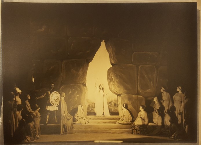 Photograph of stage production at Cornish, with a woman standing in bright light in a large crack/door in a rock wall, with men and women watching her. 