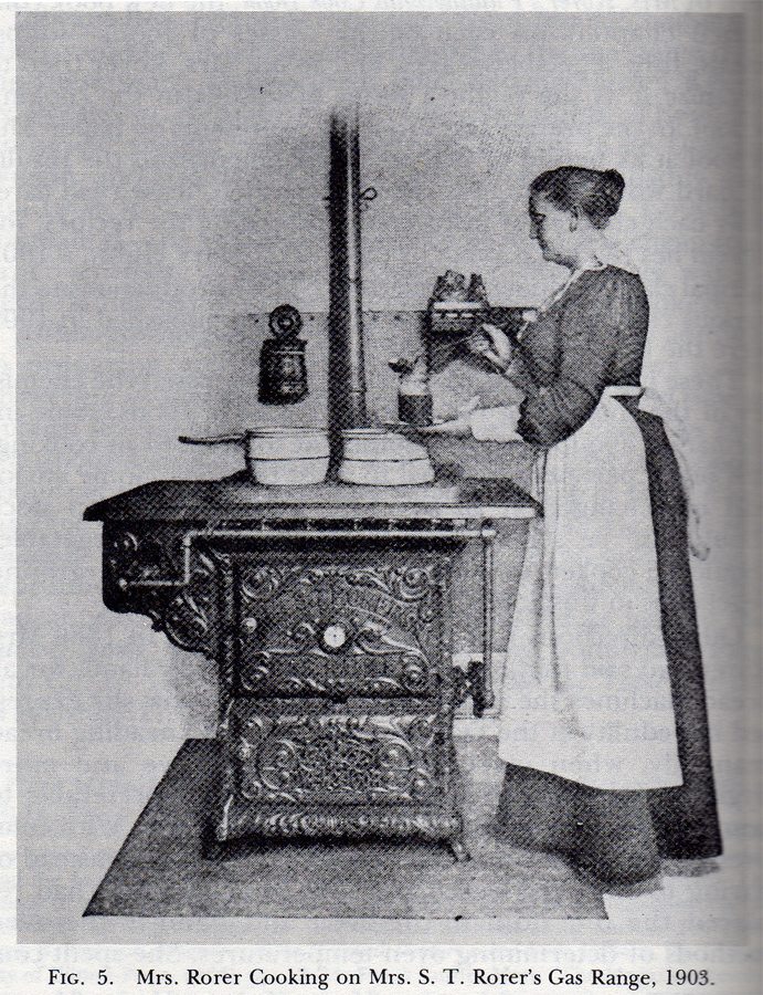 A woman in a long dress and apron standing at an early 19th century stove. 