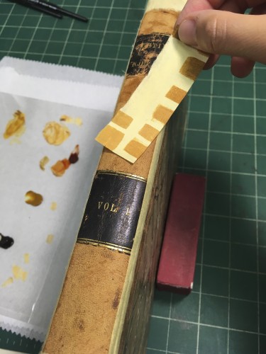 Shows several yellow-brown paint samples laid against the yellow-brown leather of the book's spine, seeking a match