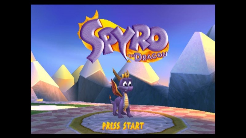 The title menu screen for the Playstation 1 game, “Spyro the Dragon”. In it stylized purple text reading “Spyro” in large letters and “The Dragon” in smaller letters are written on a circular, orange background. The majority of the image surrounding it shows a low-poly, blue-ish sky with what appear to be snow-capped mountains in the background. Beneath the title is the titular character, Spyro, who is a small purple dragon with red-orange wings, brown horns, and an orange fin on his head. The words “Press Start” are shown below Spyro in Yellow