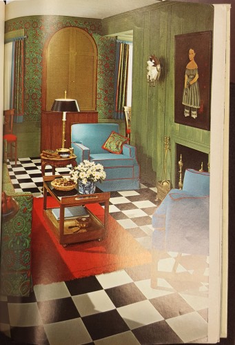 Living room with black and white tiled floor, green walls, and a green couch facing a fireplace adjacent to two blue and red chairs around a coffee table on wheels