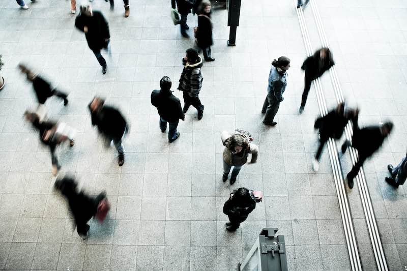 Photo taken above a busy crowd. Some figures are actively walking and their figure is blurred from their movement. Others figures are clear and sharp and they are standing, talking to or watching other people in the crowd.