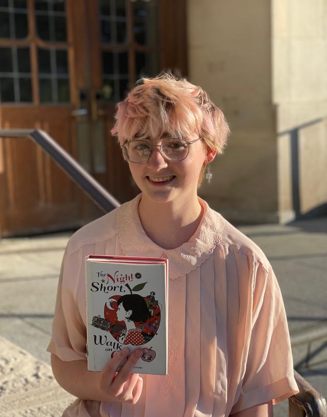 A white person with short pink hair, large round glasses, and a pink button up shirt smiles and holds up the book The Night is Short, Walk on Girl