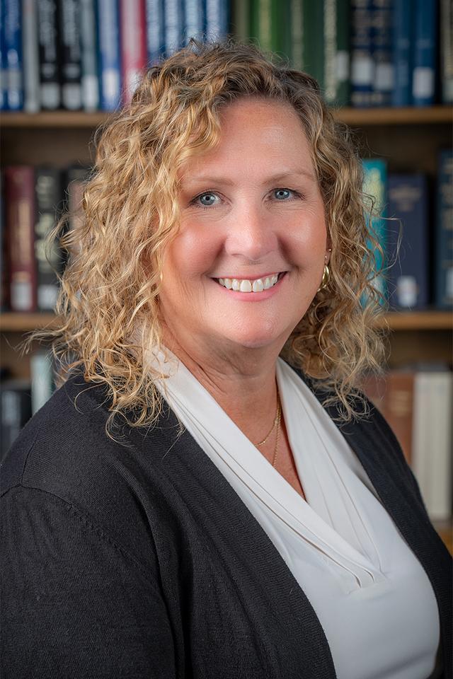 Laura Burnham, smiling woman with curly blonde hair wearing a white blouse and black cardigan, in front of a bookshelf filled with books.