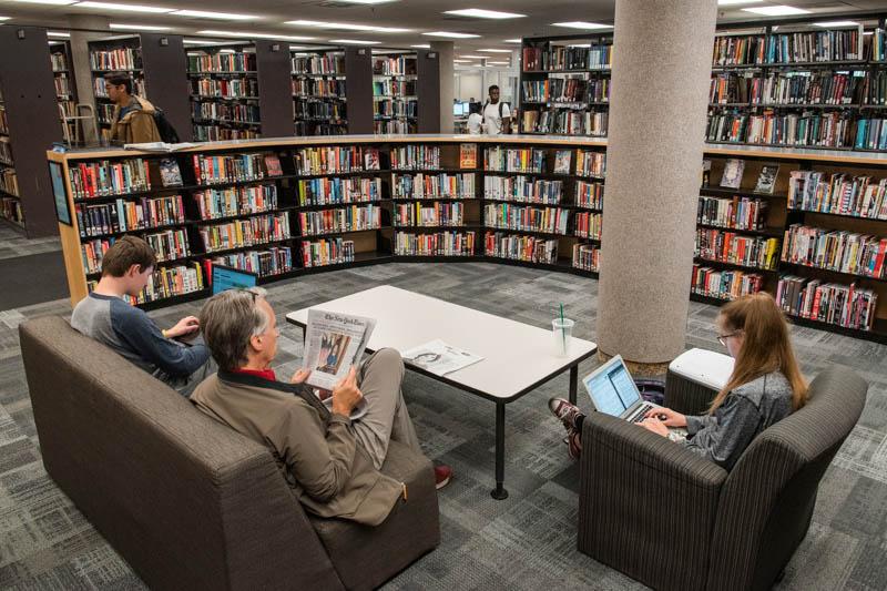 People seated while working on laptops or reading the newspaper in the Shapiro Library, with bookshelves in the background.