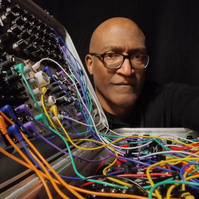 A person with glasses poses next to a modular synthesizer filled with colorful cables, set against a dark background.